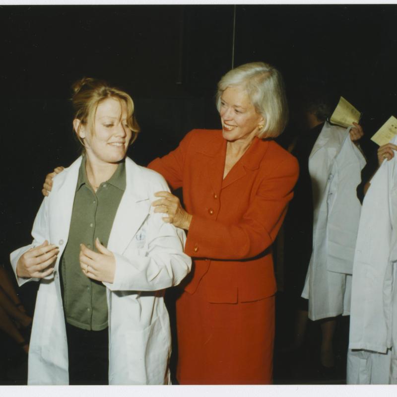 Dean Mary Mundinger assists a student into her white coat at the School of Nursing white coat ceremony.